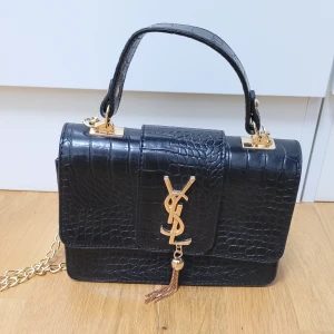 Svart YSL handväska i skinn med kedja - Säljer en svart handväska från YSL i croco-präglat skinn med guldiga detaljer och kedjerem. Väskan har ett stort YSL-emblem framtill och en dekorativ tofs. Stilren och lyxig design med både handtag och axelrem i kedja.
