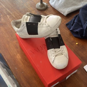 Valentino Garavani Open sneakers vit/svart - Snygga Valentino Garavani Open sneakers i vitt skinn med bred svart läderdetalj över mitten. Klassisk rund tå, vita snören och diskreta nitar på hälen. Perfekta för dig som gillar lyxiga och stilrena sneakers med en twist. Lite använda kom dm priset är inte hugget i stenen!