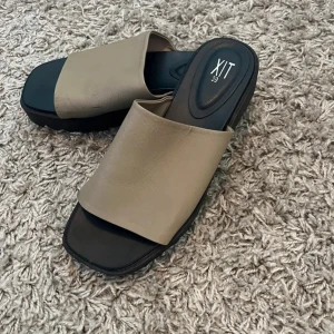 Beige sandaler från XIT med platt sula - Stilrena sandaler från XIT i beige och svart med bred rem över foten och fyrkantig tå. Platt sula och minimalistisk design, perfekta för varma dagar. Tillverkade i syntetmaterial.