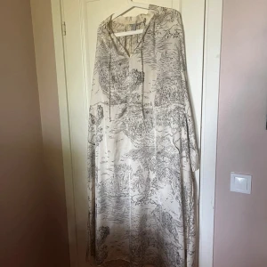 Lång vit klänning med svart mönster - Säljer en lång, luftig klänning i vitt med svart tecknat mönster. Klänningen har huva med snörning och vida ärmar, perfekt för en chill och avslappnad look. Materialet känns lätt och passar bra till varmare dagar.