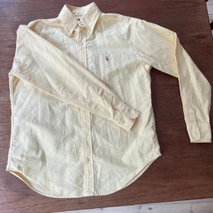 Gul skjorta från Ralph Lauren - Klassisk gul skjorta från Ralph Lauren med broderad logga på bröstet. Skjortan har lång ärm, knappar framtill och en snygg krage. Perfekt för dig som gillar stilrena och tidlösa plagg. Tillverkad i mjuk bomull för skön känsla.