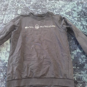 Svart Sail Racing tröja - Säljer en svart långärmad tröja från Sail Racing med vit logga tryckt på bröstet. Tröjan har rund halsringning och är tillverkad i mjuk bomull, perfekt för en chill och sportig look. Passar dig som gillar stilrena och bekväma plagg.