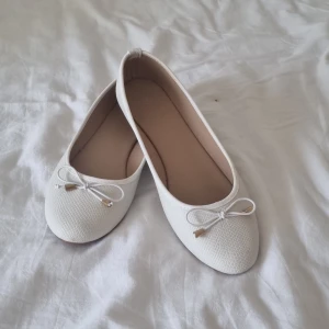 Vita ballerinaskor - Snygga vita ballerinaskor i storlek 37 med strukturmönstrad yta och söt rosett framtill. Skorna har rund tå och platt sula, vilket gör dem både stilrena och bekväma. Perfekta för dig som gillar en clean och klassisk look.