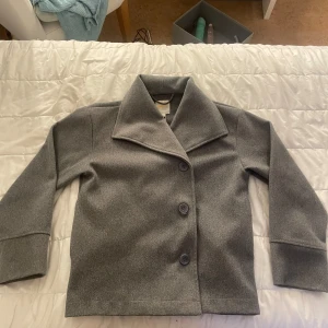 Grå kappa från Gina Tricot - Stilren grå kappa från Gina Tricot med bred krage och tre knappar framtill. Jackan har en rak passform och är tillverkad i ett mjukt ull-liknande material. Perfekt för dig som gillar en clean och klassisk look.