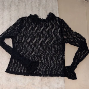 Svart spetsblus med volangkrage - Säljer en svart långärmad blus i spets med vågigt mönster. Blusen har volangdetaljer vid halsringningen och ärmsluten, vilket ger en extra snygg touch. Transparent och perfekt att styla lager på lager för en cool look.