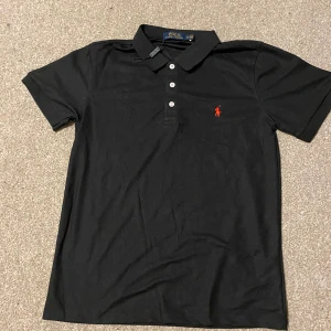 Svart pikétröja från Polo Ralph Lauren - Klassisk svart pikétröja från Polo Ralph Lauren med korta ärmar och röd broderad logga på bröstet. Tröjan har krage och tre knappar framtill. Tillverkad i mjuk bomull som känns skön mot huden.