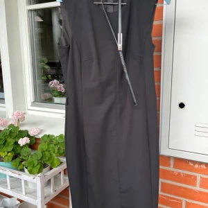 Svart ärmlös långklänning med bälte - Stilren svart långklänning med ärmlös design och medföljande smalt bälte i midjan. Klänningen har en rak och enkel siluett som ger en klassisk look. Perfekt för dig som gillar minimalistisk stil och vill ha något tidlöst i garderoben. 