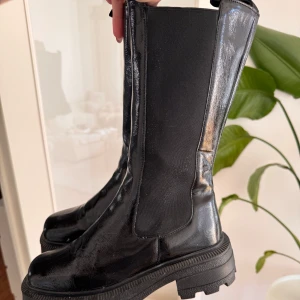 Svarta glansiga chelsea boots - Snygga svarta chelsea boots med glansig finish från Nelly. Skorna har elastiska paneler på sidorna för enkel påtagning och en chunky, platt sula som ger en cool look. Perfekta till både jeans och kjol/klänning. Endast använt en gång är i jättebra skick