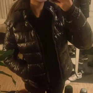 Svart pufferjacka från 157 - Snygg svart pufferjacka med glansig finish från 157. Jackan har en hög krage, dragkedja framtill och två sidofickor. Perfekt för kalla dagar och ger en cool vibe till din outfit. Passformen är normal och jackan är vadderad för extra värme.