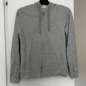 Ljusgrå hoodie med dragkedja från Lager157 - Enkel och stilren ljusgrå hoodie från Lager 157. Hoodien är i ljusare grå färg än vad bilden visar. Den har dragkedja framtill, huva med snörning och två fickor. Storlek M, men passar även om man har storlek S. Aldrig använd. 