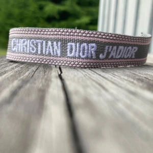 Christian Dior J'Adior armband - Superfint armband från Christian Dior med texten 'J'Adior' broderad i vitt på grå och ljusrosa bakgrund. Armbandet är i tyg med justerbar knytning och tofsar i ändarna. Perfekt accessoar för att lyfta din outfit med en lyxig touch. Priset kan förhandlas, hör av er vid frågor❤️🤝