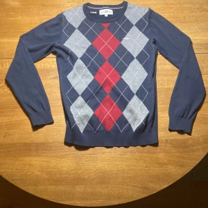 Argyletröja Hampton Republic blå/röd - Klassisk långärmad tröja från Hampton Republic i marinblått med argylemönster i grått, rött och blått. Tröjan är i 100% bomull och har ribbade muddar vid ärmslut och nederkant. Perfekt för dig som gillar preppy stil.