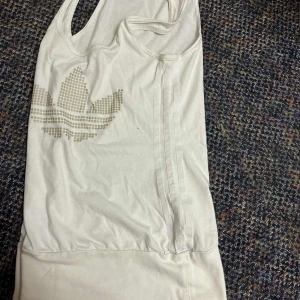 Vit linnetopp - Säljer en vit linnetopp från Adidas med det klassiska Trefoil-loggan i beige prickigt mönster framtill. Har strecken på sidorna. 🩷