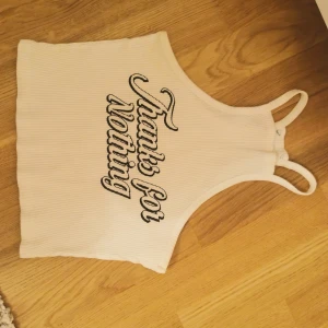 Vit ribbad croppad linnetopp - Säljer en vit ribbad croppad linnetopp med svarta bokstäver framtill där det står 'Thanks for Nothing'. Toppen har smala axelband och är ärmlös, perfekt för en chill och avslappnad stil.