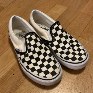 Vans slip-on checkerboard sneakers - Klassiska Vans slip-on sneakers med svartvitt rutigt mönster. Skorna har låg profil, vit sula och är tillverkade i canvas. Perfekta för en avslappnad och cool streetstil.