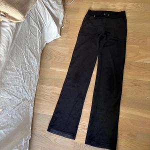 Svarta velourbyxor Juicy Couture XXS - Svarta mjukisbyxor från Juicy Couture i velour med broderad JC-logga framtill. Byxorna har snörning i midjan, raka ben och fickor både fram och bak med silvriga knappar. Perfekta för en chill och trendig look. Har follat upp byxorna innan och sedan har follen lossnat, men den är enkel att få bort