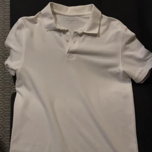 Vit pikétröja från Dressmann - Klassisk vit pikétröja från Dressmann i storlek L. Tröjan har korta ärmar, krage och knäppning med två knappar framtill. Perfekt basic-plagg med stilren look, tillverkad i mjuk bomull som känns skön mot huden.