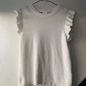Vit stickad topp - Supersöt vit ribbad topp från Vila med volangdetaljer vid axlarna. Den är ärmlös och har en rund halsringning. Materialet är mjukt och stickat.