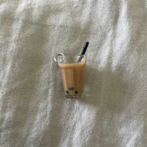 Bubble tea-berlock i miniatyr. - Supergulligt halsband med en miniatyr av ett glas bubble tea, komplett med svart sugrör och små svarta pärlor som boba. Hängsmycket är i plast och har en beige och svart färgskala. Perfekt accessoar för dig som älskar bubble tea och vill sticka ut.