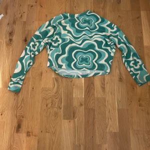 Grön och vit mönstrad långärmad tröja H&M - Säljer en ribbad långärmad tröja från H&M i storlek 146/152. Tröjan har ett coolt grönt och vitt vågigt mönster med blommiga detaljer och rund hals. Perfekt för dig som gillar färg och retro vibes!
