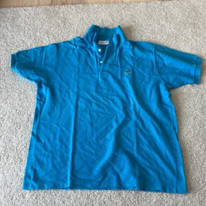 Blå pikétröja från Lacoste XL - Snygg blå pikétröja från Lacoste i klassisk modell med krage och två knappar framtill. Ikoniska krokodilloggan på bröstet. Tröjan är i bomull och har korta ärmar, perfekt för sommaren. Passar dig som gillar stilren och sportig vibe.