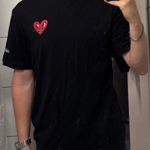 Svart t-shirt med rött hjärta-tryck - Svart t-shirt från HM X Keith Haring med ett stort rött hjärta med ansikte tryckt på ryggen och ett mindre hjärta på bröstet. Klassisk passform och rund hals. Materialet är mjuk bomull, perfekt för en chill och stilren look.
