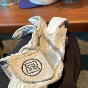 Vita golfhandskar från FootJoy - Snygga vita golfhandskar från FootJoy med diskreta grå detaljer och perforeringar för extra ventilation. Tillverkade i mjukt skinn med kardborreknäppning och broderad logga på handleden. Perfekt grepp och stilren look på golfbanan. den är bäldigt fet