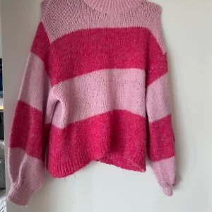 Randig stickad tröja från Gina Tricot - Supermysig stickad tröja från Gina Tricot i breda rosa och ceriseröda ränder. Tröjan har en hög, ribbad hals och långa ärmar med ribbade muddar. Perfekt för dig som gillar färg och vill ha en chill oversized vibe. Den är tyvärr lite nopprig och har tecken på slitage men fortfarande sjukt fin
