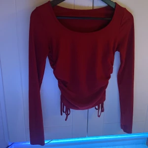 Röd långärmad topp med snörning - Trendig röd långärmad topp med rund halsringning och snygg snörning i sidorna som ger en cool rynkad effekt. Perfekt för dig som vill ha en enkel men ändå unik look. Toppens material är ribbat och känns mjukt mot huden.