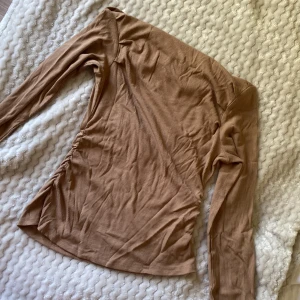 Beige ribbad offshoulder topp - Säljer en beige ribbad offshoulder topp med lång ärm och snygg rynkdetalj på sidan. Toppens material är mjukt och stretchigt, perfekt för en trendig och avslappnad look. Passformen är slim och toppen är lätt croppad för en modern vibe. Endast använt en gång.