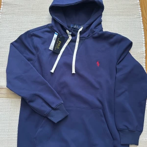 Marinblå hoodie från Polo Ralph Lauren - Snygg marinblå hoodie från Polo Ralph Lauren med vit snörning och klassisk röd logga broderad på bröstet. Tröjan har huva, känguruficka och ribbade muddar. Perfekt för en chill och stilren look. Säljes för 635kr, aldrig används sälja för den inte kommer till användning.