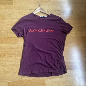 Vinröd t-shirt från Calvin Klein Jeans - Snygg vinröd t-shirt från Calvin Klein Jeans med tryckt logga i orange på bröstet. Klassisk rund halsringning och korta ärmar. Skön passform i mjuk bomull, perfekt till jeans eller shorts.