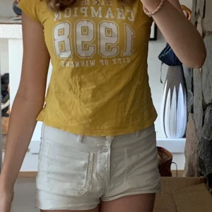 Gul t-shirt Champion 1998 H&M - Gul t-shirt från H&M med trycket 'Champion 1998' i vitt framtill. Modellen är kortärmad och har en klassisk rund halsringning. T-shirten är i mjuk bomull och har en avslappnad passform som är perfekt till jeans eller shorts.