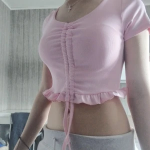 (1st 40kr) Rosa croppad topp med volangkant - Supersöt ljusrosa croppad topp med korta ärmar, ribbad struktur och volangkant nertill. Toppen har snörning framtill som ger en snygg rynkad effekt. Perfekt för dig som gillar gulliga och trendiga plagg.