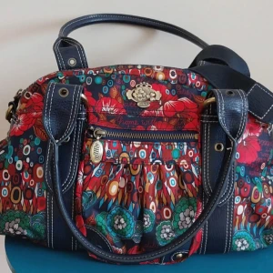 Oilily handväska  - Superfin handväska från Oilily med blommigt och färgstarkt mönster i rött, grönt, blått och vitt. Väskan har marinblå detaljer, dubbla handtag, justerbar axelrem och dragkedja. Skicket är väldigt bra men kan ha något mycket litet slitage som inte märks av. Långt band kommer också med. Skriv gärna om ni har några frågor!!