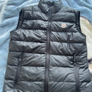Svart dunväst från Moncler - Snygg svart dunväst från Moncler med klassisk quiltad design och hög krage. Västen har två dragkedjefickor framtill och Monclers ikoniska logga på bröstet. Perfekt att slänga över en hoodie eller tröja för extra värme och stil.