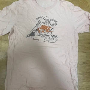 Ljusrosa Nike t-shirt med tryck - Säljer en ljusrosa t-shirt från Nike med coolt tryck av palmer och en hängmatta på bröstet. T-shirten är i mjuk bomull och har klassisk passform med korta ärmar. Perfekt för dig som gillar avslappnad stil och streetwear.