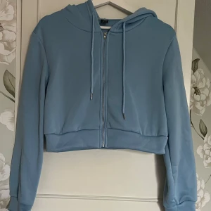 Ljusblå croppad hoodie med dragkedja - Säljer en ljusblå croppad hoodie med dragkedja framtill och snörning i huvan. Tröjan har långa ärmar och en skön, mjuk känsla.