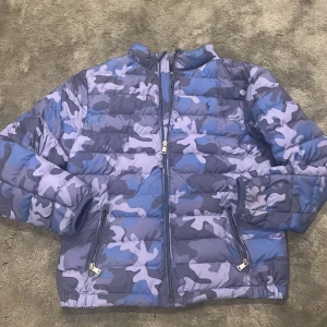 Camouflage pufferjacka från Polo Ralph Lauren - Säljer en fet pufferjacka från Polo Ralph Lauren med blått och grått camouflage-mönster. Jackan har dragkedja framtill, två fickor med dragkedja och klassisk Polo-logga på bröstet. Perfekt för kalla dagar när du vill sticka ut lite extra.