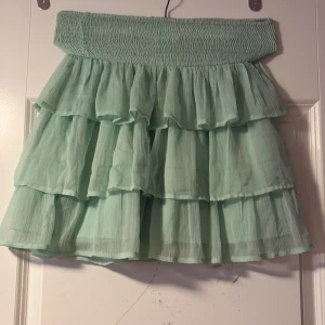 Mintgrön volangkjol med resårmidja - Superfin mintgrön kortkjol med flera lager volanger och bred smockresår i midjan. Kjolen är luftig och lätt, perfekt för varma dagar. Ger en söt och trendig look med sin ljusa färg och flowiga form.