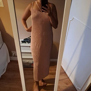 Plisserad champagnefärgad långklänning - Säljer en elegant plisserad långklänning i champagnefärg med smala axelband och rak passform. Klänningen har en stilren look och är perfekt för dig som gillar minimalistisk stil och vill sticka ut med en mjuk, glansig ton.