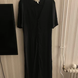 Svart långklänning med knappar - Stilren svart långklänning med v-ringning och klädda knappar framtill. Klänningen har korta ärmar och är tillverkad i ett mjukt, lätt material som faller snyggt. Perfekt för dig som gillar minimalistisk och tidlös stil.