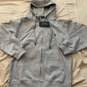 Grå hoodie från Polo Ralph Lauren - Snygg grå hoodie från Polo Ralph Lauren med dragkedja och huva. Klassisk logga broderad på bröstet, ribbade muddar och känguruficka framtill. Perfekt för chill dagar och enkel att matcha med jeans eller joggers.