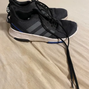 Svarta Adidas sneakers med Cloudfoam - Svarta Adidas sneakers med mesh-ovandel och klassiska tre ränder i grått på sidorna. Skorna har vit sula med blå och svarta detaljer samt Cloudfoam-dämpning för extra komfort. Perfekta för dig som gillar sportig och stilren look.