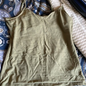 Olivgrön linnetopp med smala axelband - Enkel och stilren olivgrön linnetopp med smala axelband och v-ringad hals. Toppen är ärmlös och har en rak passform, perfekt att matcha med jeans eller kjol. Mjuk och lätt i materialet, vilket gör den skön att bära under varma dagar.
