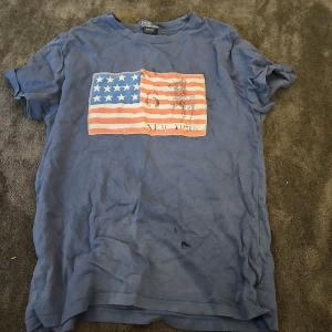 Blå t-shirt Polo Ralph Lauren med flagga - Blå t-shirt från Polo Ralph Lauren med tryck av amerikanska flaggan på bröstet. Klassisk rund hals och korta ärmar. Skön och mjuk bomull, perfekt för en avslappnad stil.