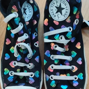 Svarta sneakers med hjärtan - Coola svarta sneakers med färgglada, holografiska hjärtan över hela skon. Klassisk vit gummitå och sula, samt vita snören. Perfekta för dig som vill sticka ut med ett lekfullt mönster och streetstyle-vibe.