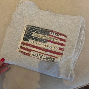 Grå hoodie Ralph Lauren Denim & Supply - Snygg grå hoodie från Ralph Lauren Denim & Supply med amerikansk flagga och vintage-tryck på bröstet. Tröjan har huva, magficka och långa ärmar. Perfekt för dig som gillar streetstyle och vill ha en chill look. Lite missfärgad på lapp och lite i luvan. Annars superskick