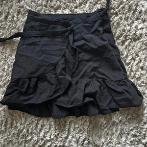 Svart omlottkjol med volang NLY Trend - Svart kort omlottkjol från NLY Trend i storlek 32. Kjolen har volangkant nertill och knyts i midjan för en snygg passform. Perfekt för dig som gillar en enkel men trendig look.