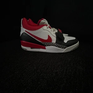 Nike Jordan 1 Low vit/röd/svart - Nike Jordan 1 Low sneakers i vitt, rött och svart med klassisk röd swoosh och svart rem med Nike-logga. Skorna har perforerad tå, grå och röd yttersula samt ikonisk Jumpman-logga på hälen. Ovandel i skinn och textil med detaljer och mönstrad panel vid sulan.